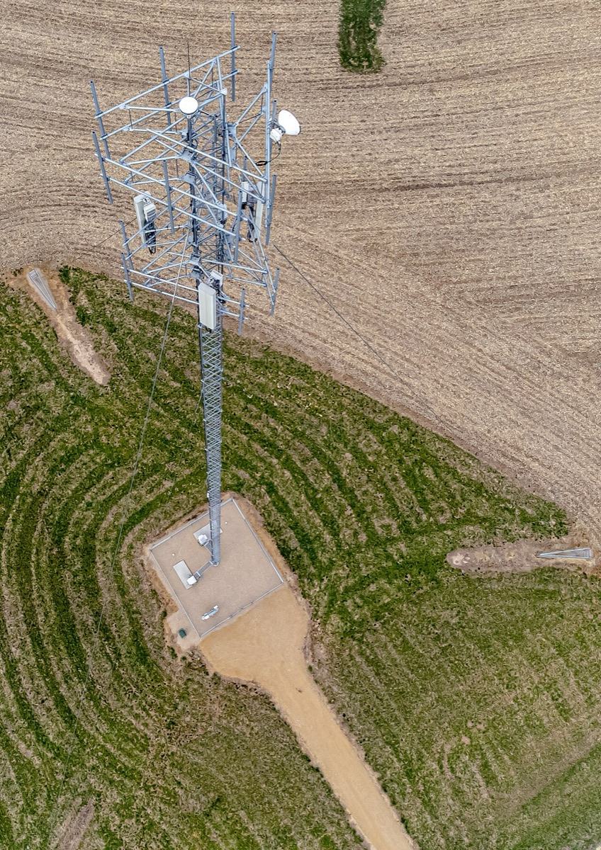 Aerial view of a lattice cell tower with antennas on a small gravel lease pad in a farm field
